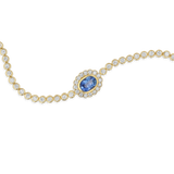 A gold chain bracelet featuring a central oval blue sapphire stone surrounded by a halo of small clear stones, with the rest of the chain lined with round clear stones, shown on a plain white background.