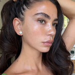 A close-up portrait of a woman with long, dark brown hair, wearing a large silver stud earring and a green strap top, with her hand pulling her hair back.