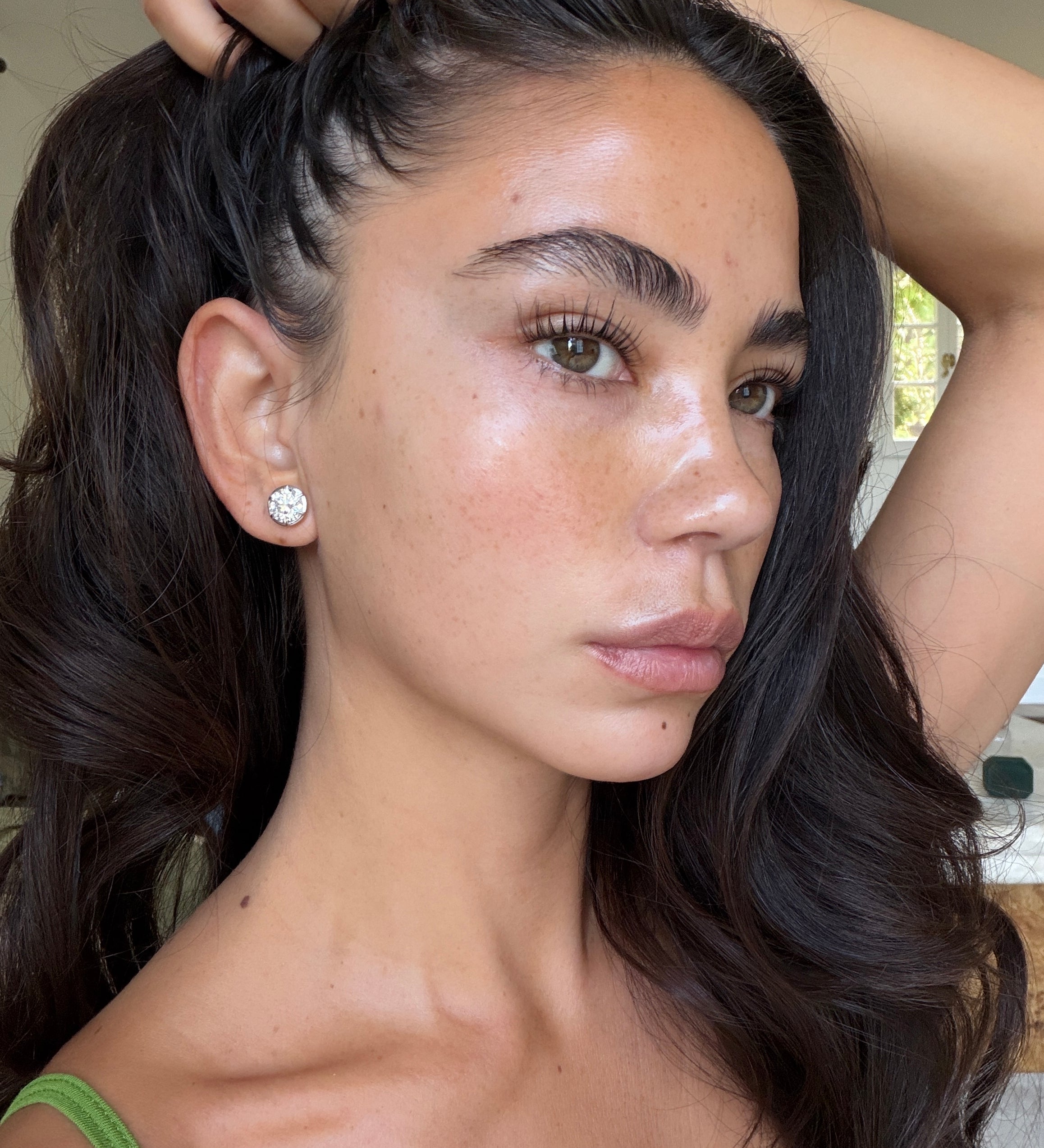 A close-up portrait of a woman with long, dark brown hair, wearing a large silver stud earring and a green strap top, with her hand pulling her hair back.