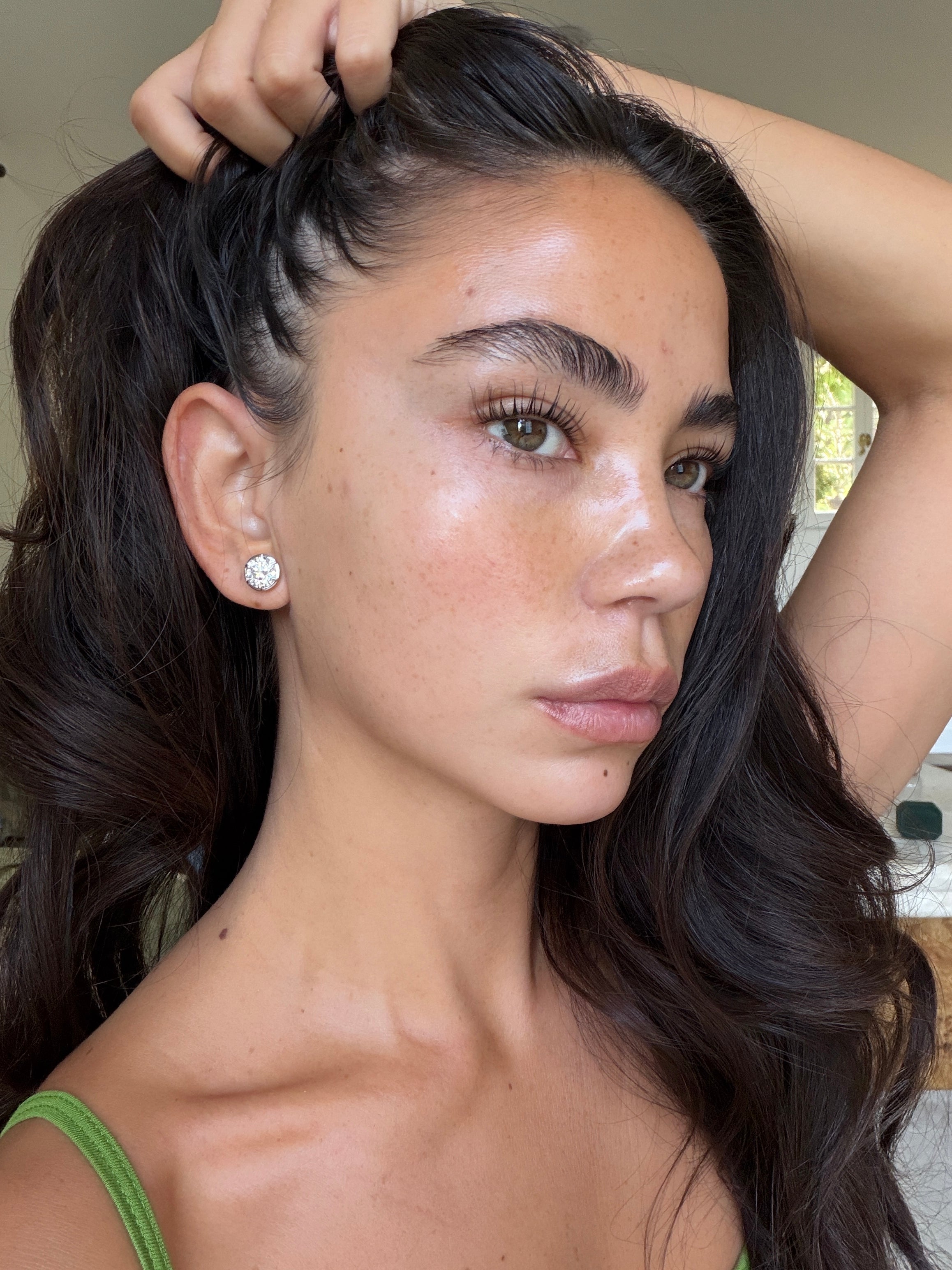 A close-up portrait of a woman with long, dark brown hair, wearing a large silver stud earring and a green strap top, with her hand pulling her hair back.