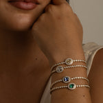 A close-up of a woman's face and hand resting near her neck, showcasing three delicate silver-toned beaded bracelets with round bezel-set stones in silver, blue, and green on her wrist.