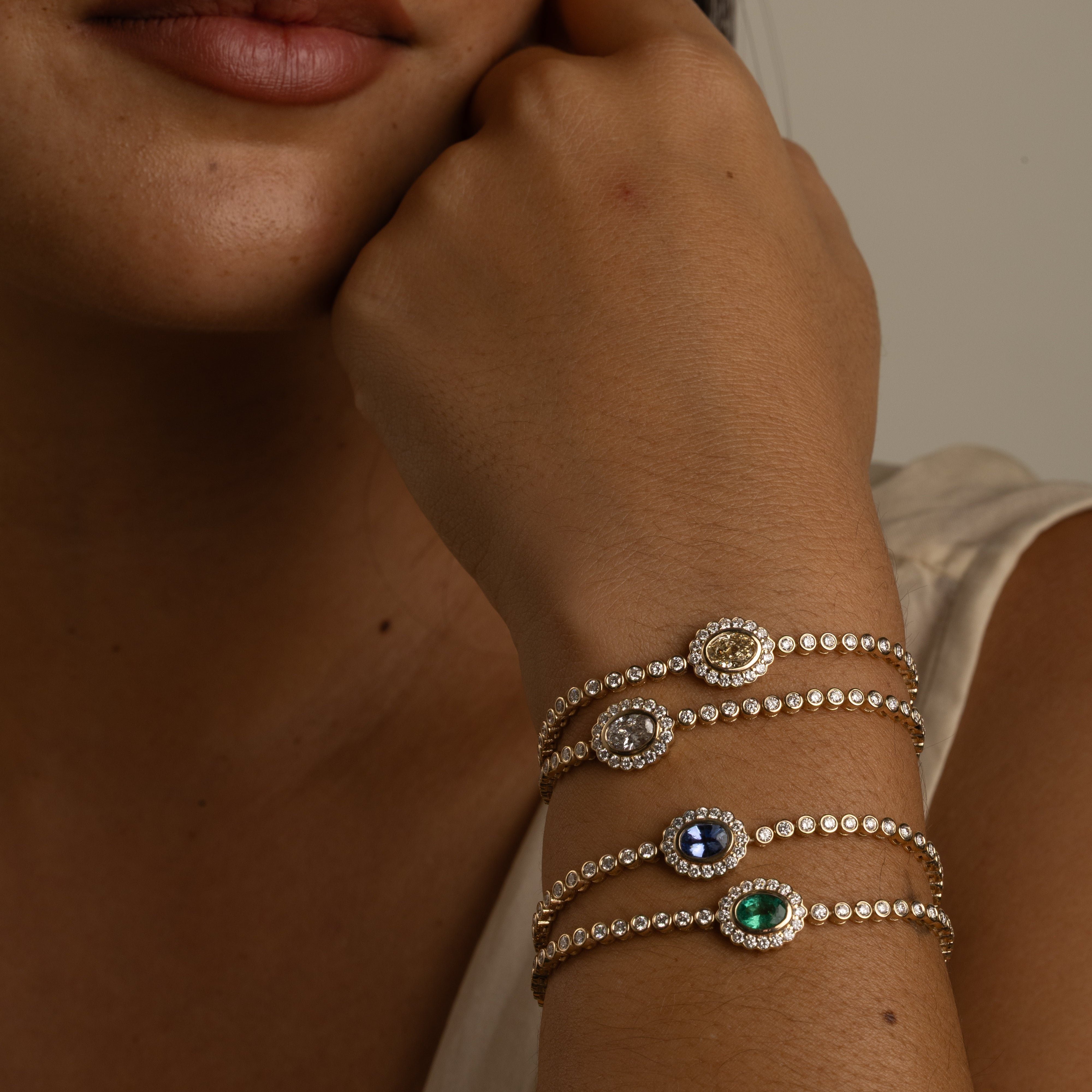 A close-up of a woman's face and hand resting near her neck, showcasing three delicate silver-toned beaded bracelets with round bezel-set stones in silver, blue, and green on her wrist.
