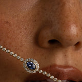 A close-up of a model's face wearing a silver-toned chain necklace with a central oval blue gemstone surrounded by small clear stones.