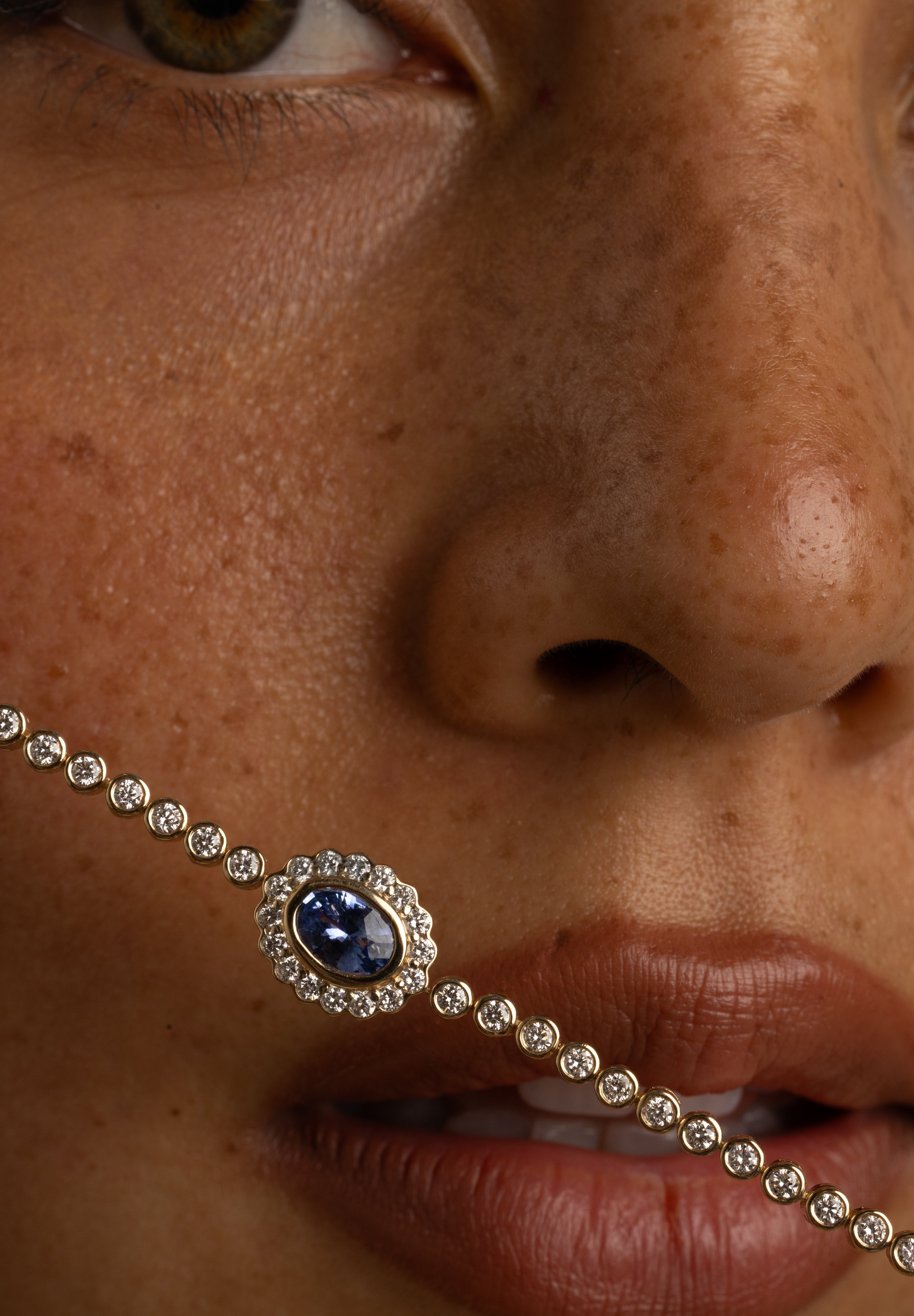 A close-up of a model's face wearing a silver-toned chain necklace with a central oval blue gemstone surrounded by small clear stones.