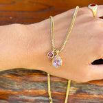 A gold chain necklace with a teardrop-shaped light purple pendant and a small round purple gemstone, shown worn on a person's wrist against a rustic wooden background.