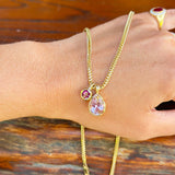 A gold chain necklace with a teardrop-shaped light purple pendant and a small round purple gemstone, shown worn on a person's wrist against a rustic wooden background.