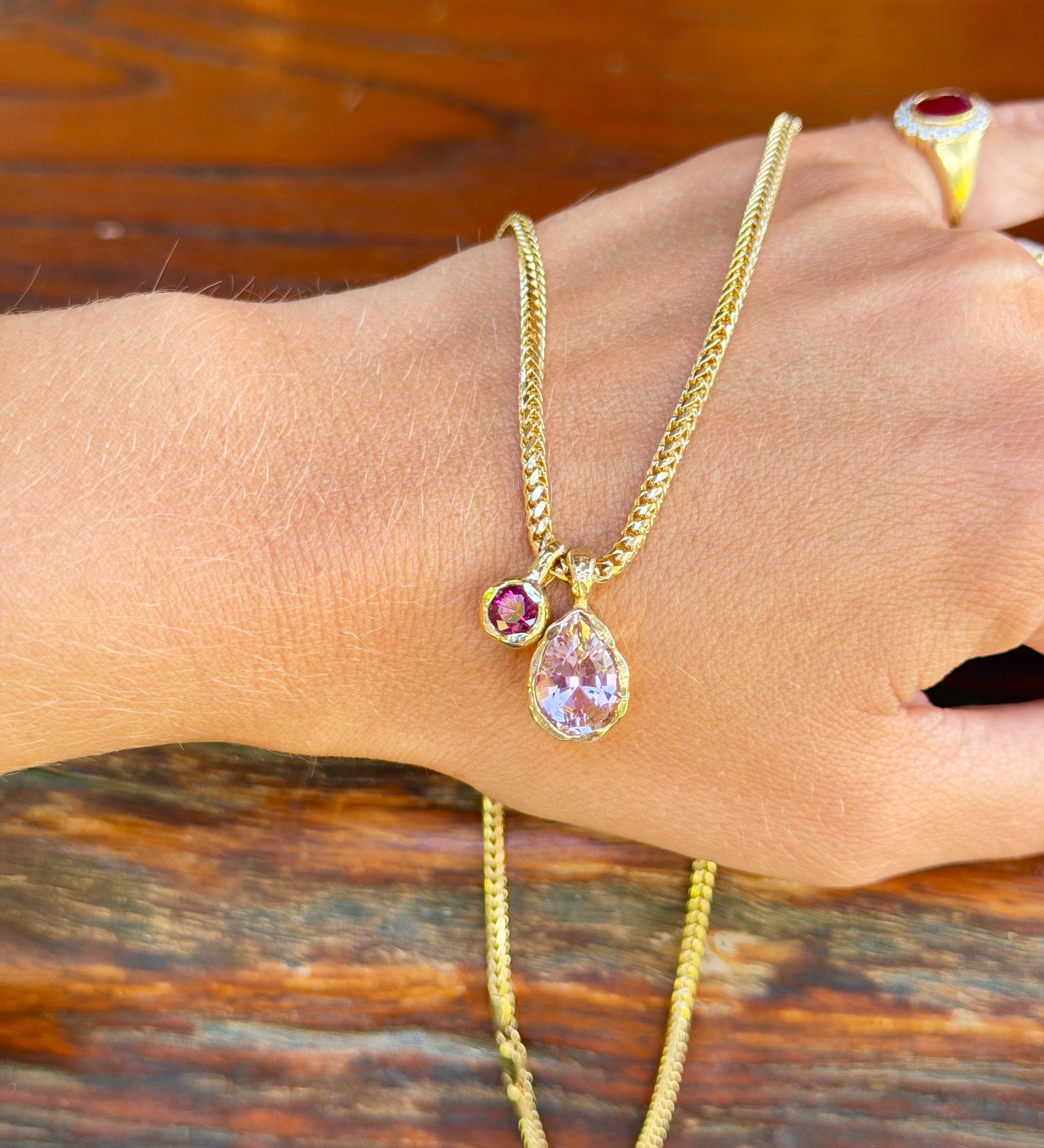 A gold chain necklace with a teardrop-shaped light purple pendant and a small round purple gemstone, shown worn on a person's wrist against a rustic wooden background.