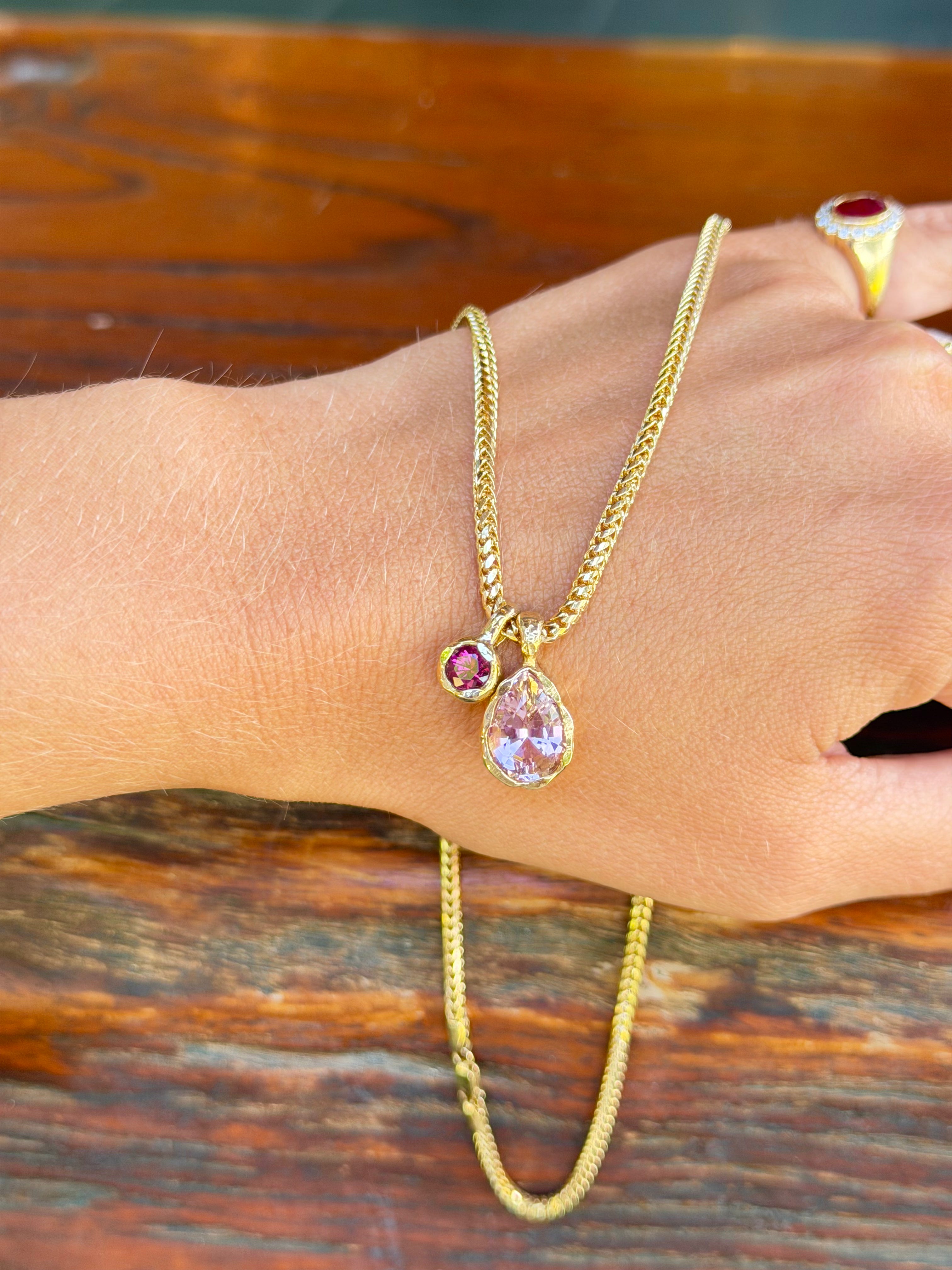 A gold chain necklace with a teardrop-shaped light purple pendant and a small round purple gemstone, shown worn on a person's wrist against a rustic wooden background.