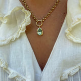 A gold chain necklace with a teardrop-shaped green gemstone pendant, worn by a person in a white ruffled blouse.