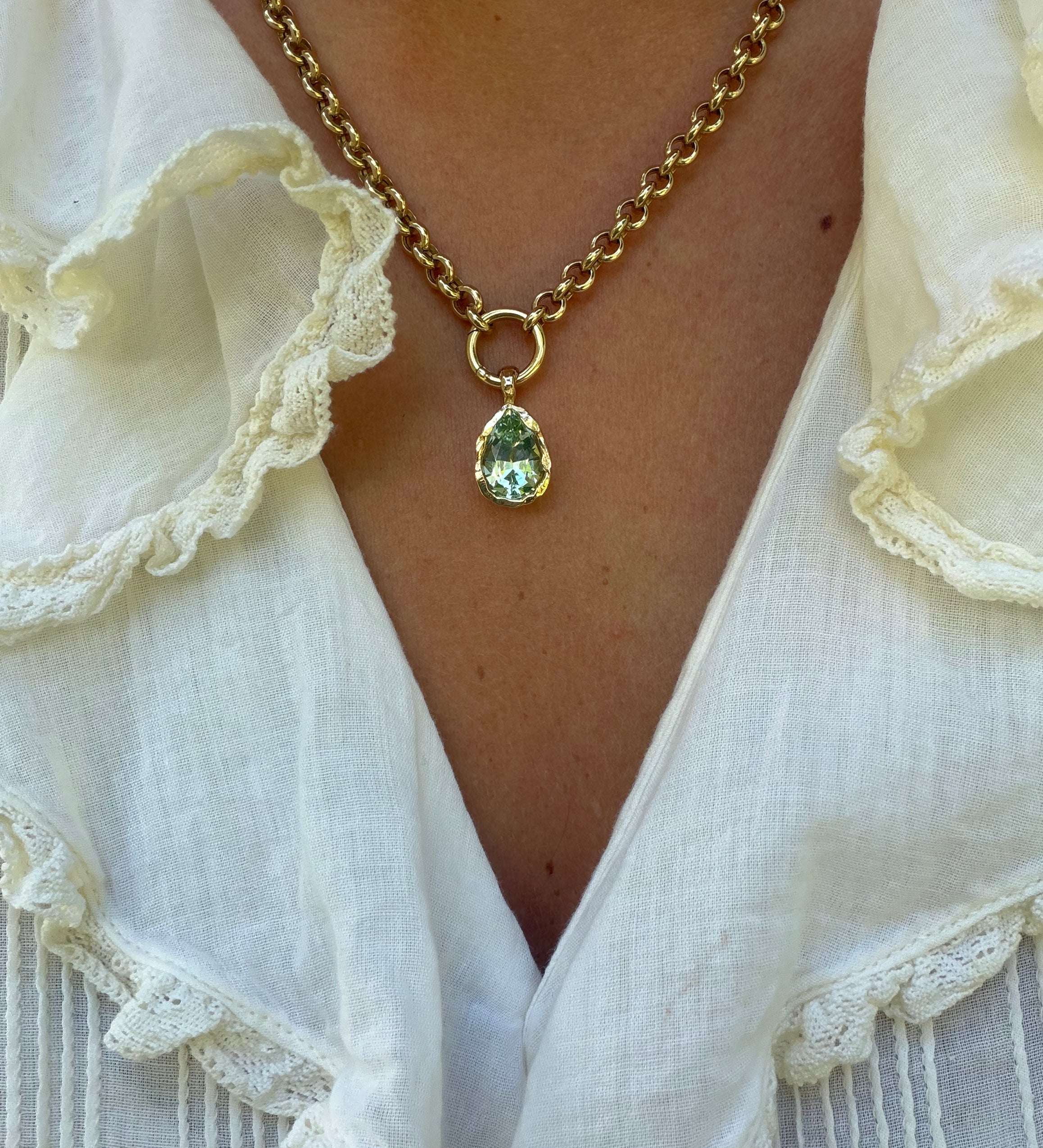 A gold chain necklace with a teardrop-shaped green gemstone pendant, worn by a person in a white ruffled blouse.