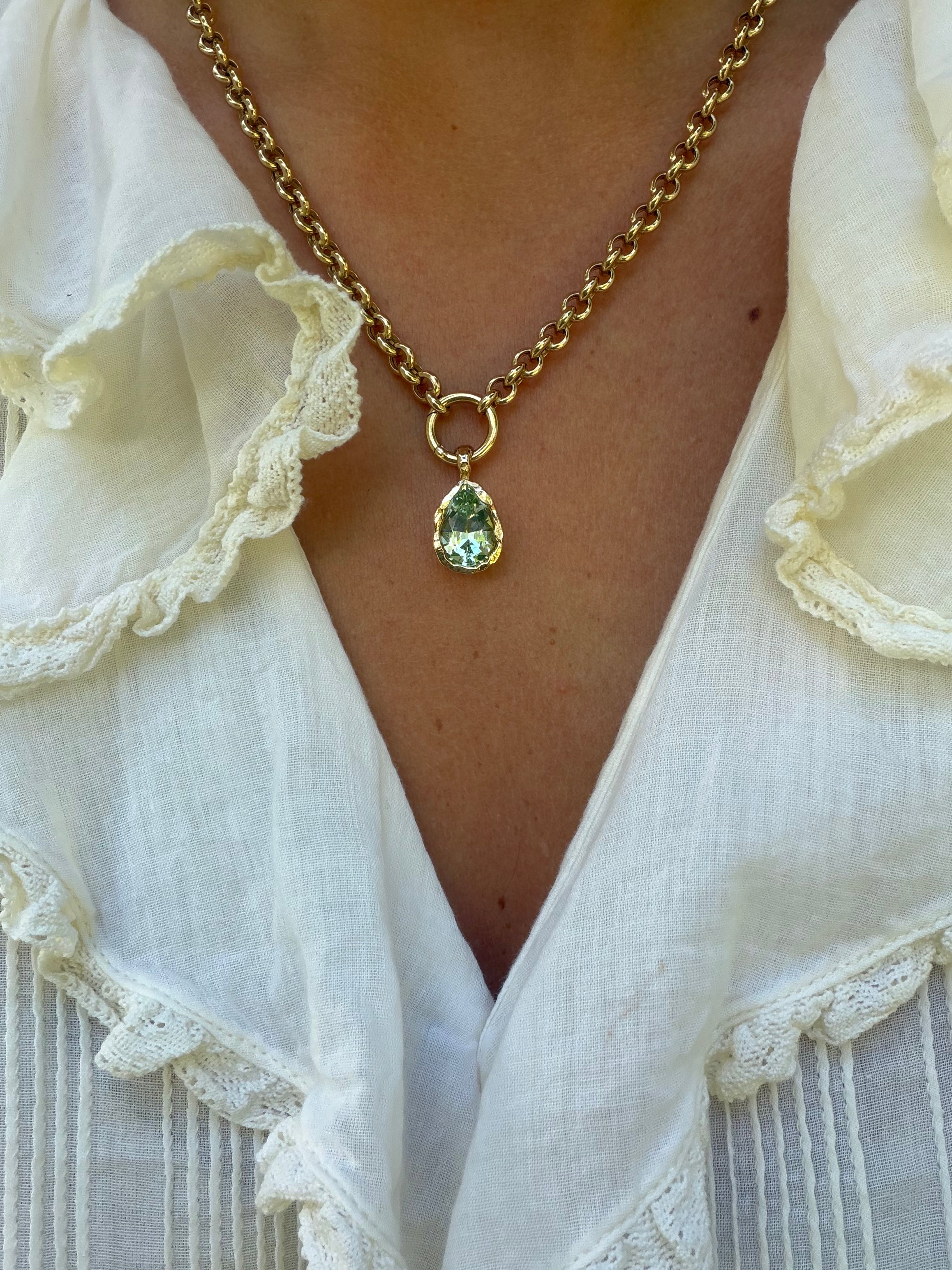 A gold chain necklace with a teardrop-shaped green gemstone pendant, worn by a person in a white ruffled blouse.