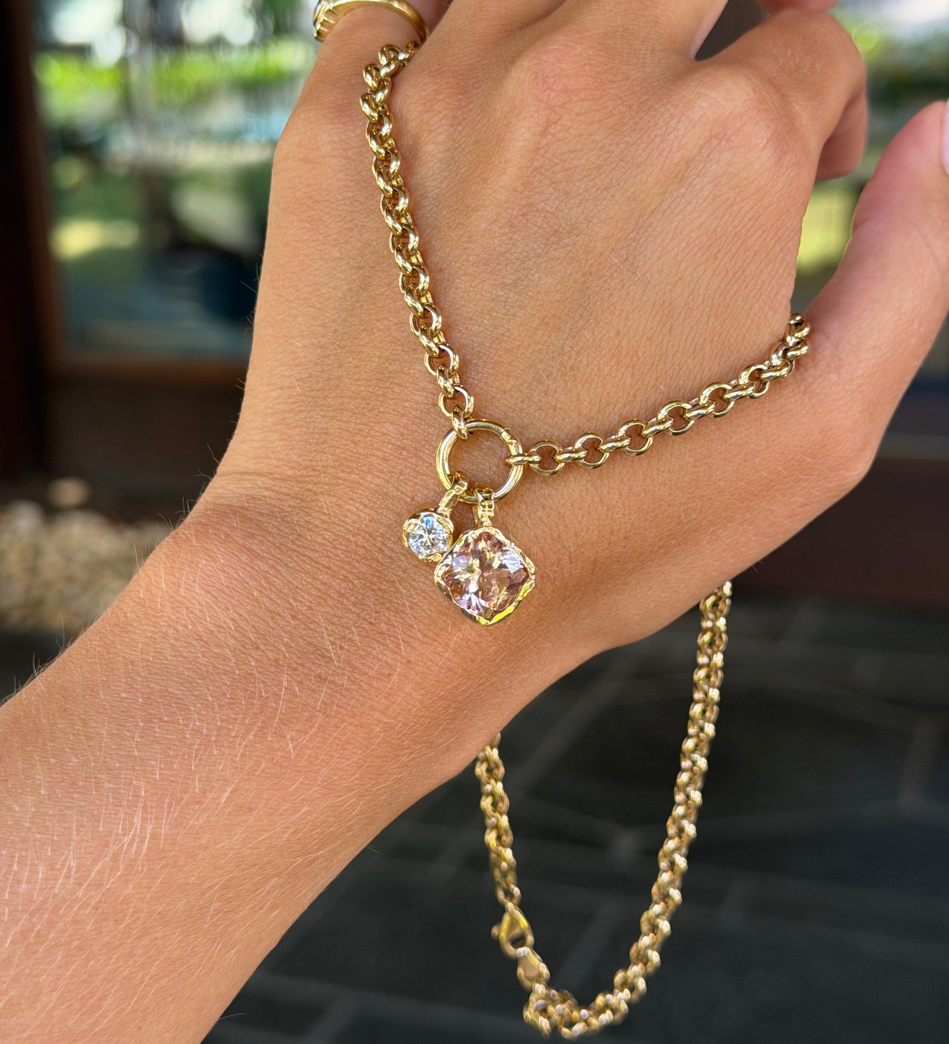 A gold chain hand bracelet with a small pendant featuring a clear faceted gemstone, shown worn on a person's hand with gold rings on the fingers.