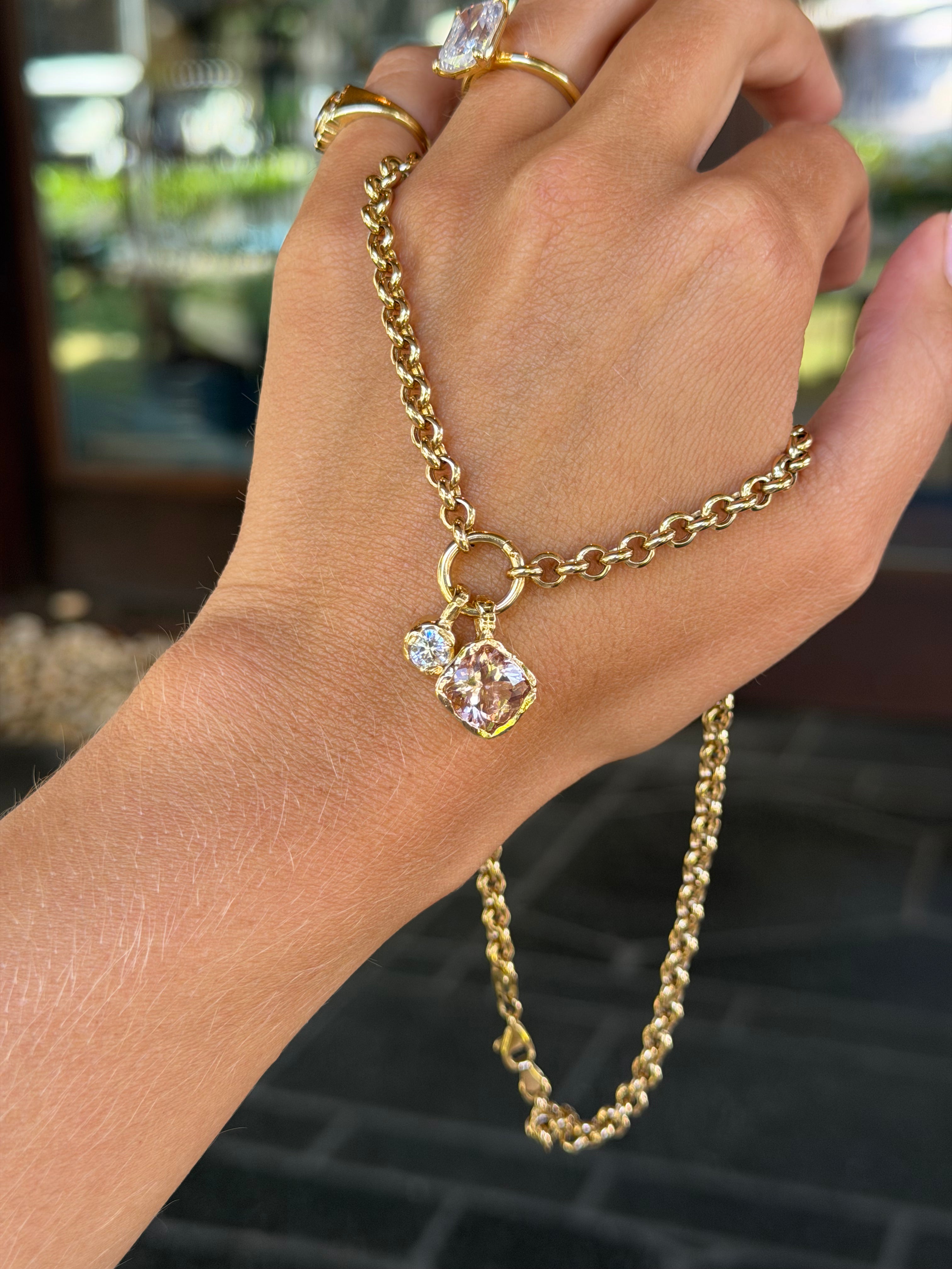 A gold chain hand bracelet with a small pendant featuring a clear faceted gemstone, shown worn on a person's hand with gold rings on the fingers.