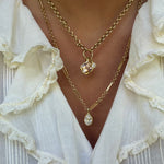 A close-up of a person wearing layered gold necklaces over a white ruffled blouse. One necklace has a small rectangular pendant with a clear faceted stone, and the other has a teardrop-shaped clear pendant.