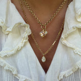 A close-up of a person wearing layered gold necklaces over a white ruffled blouse. One necklace has a small rectangular pendant with a clear faceted stone, and the other has a teardrop-shaped clear pendant.