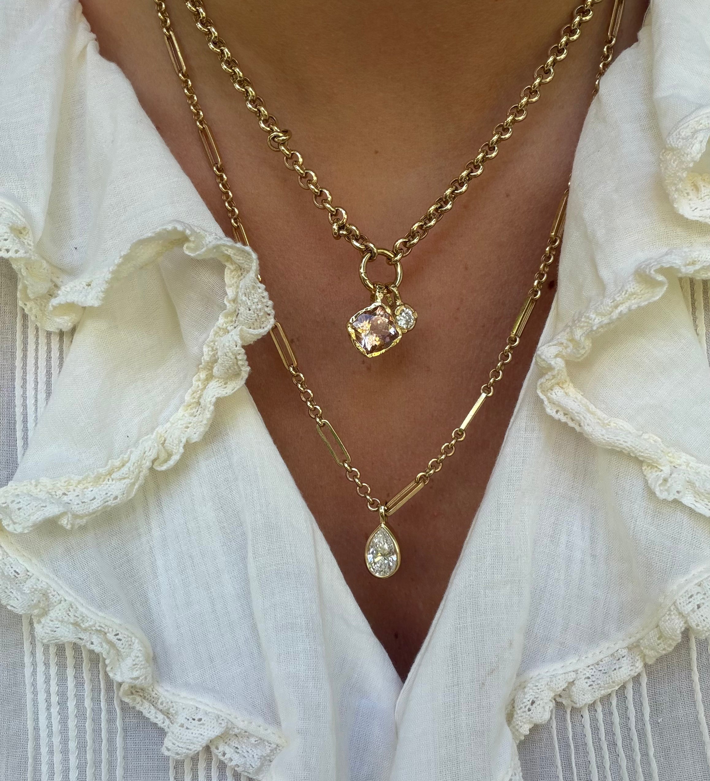 A close-up of a person wearing layered gold necklaces over a white ruffled blouse. One necklace has a small rectangular pendant with a clear faceted stone, and the other has a teardrop-shaped clear pendant.