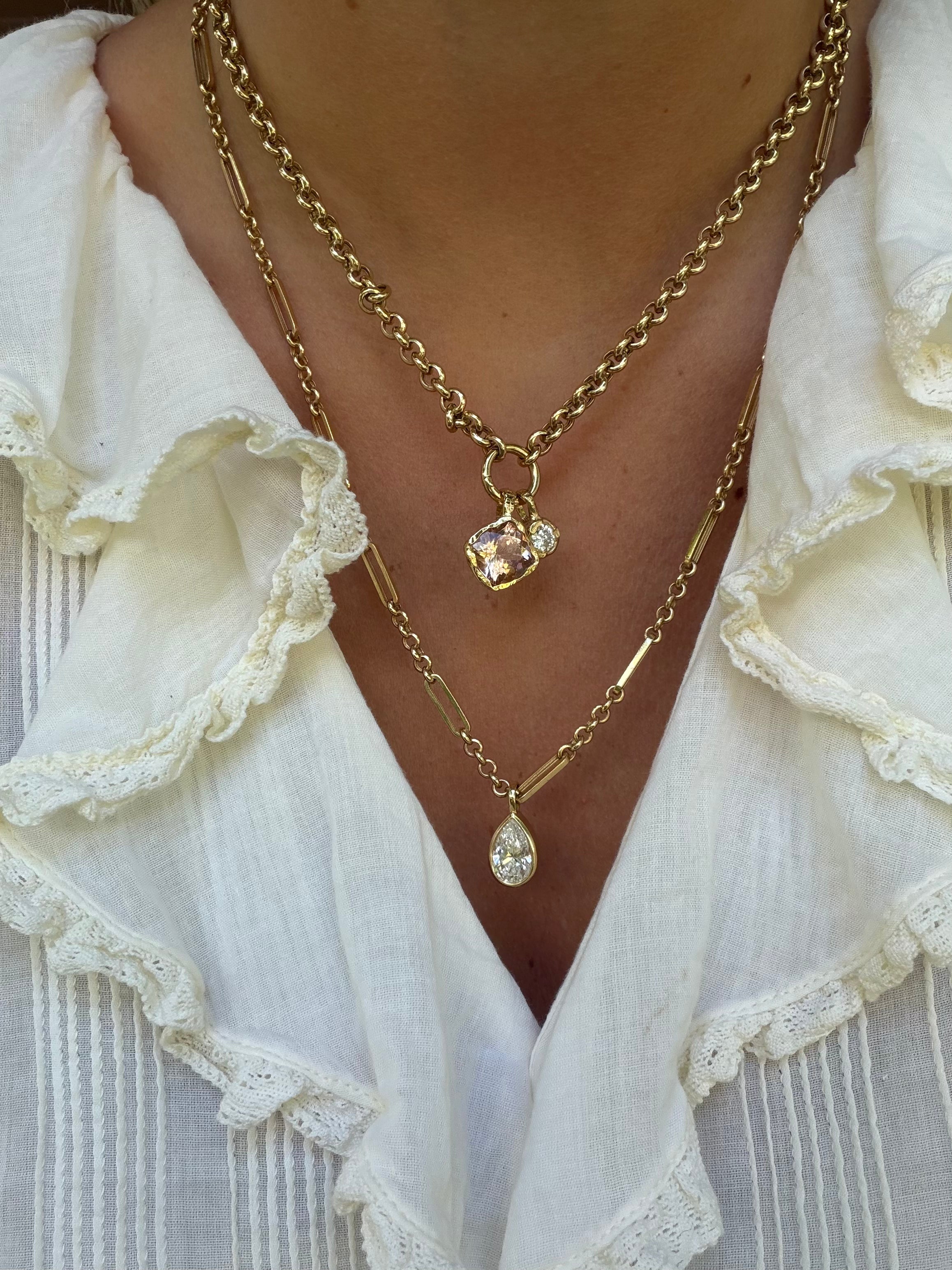 A close-up of a person wearing layered gold necklaces over a white ruffled blouse. One necklace has a small rectangular pendant with a clear faceted stone, and the other has a teardrop-shaped clear pendant.