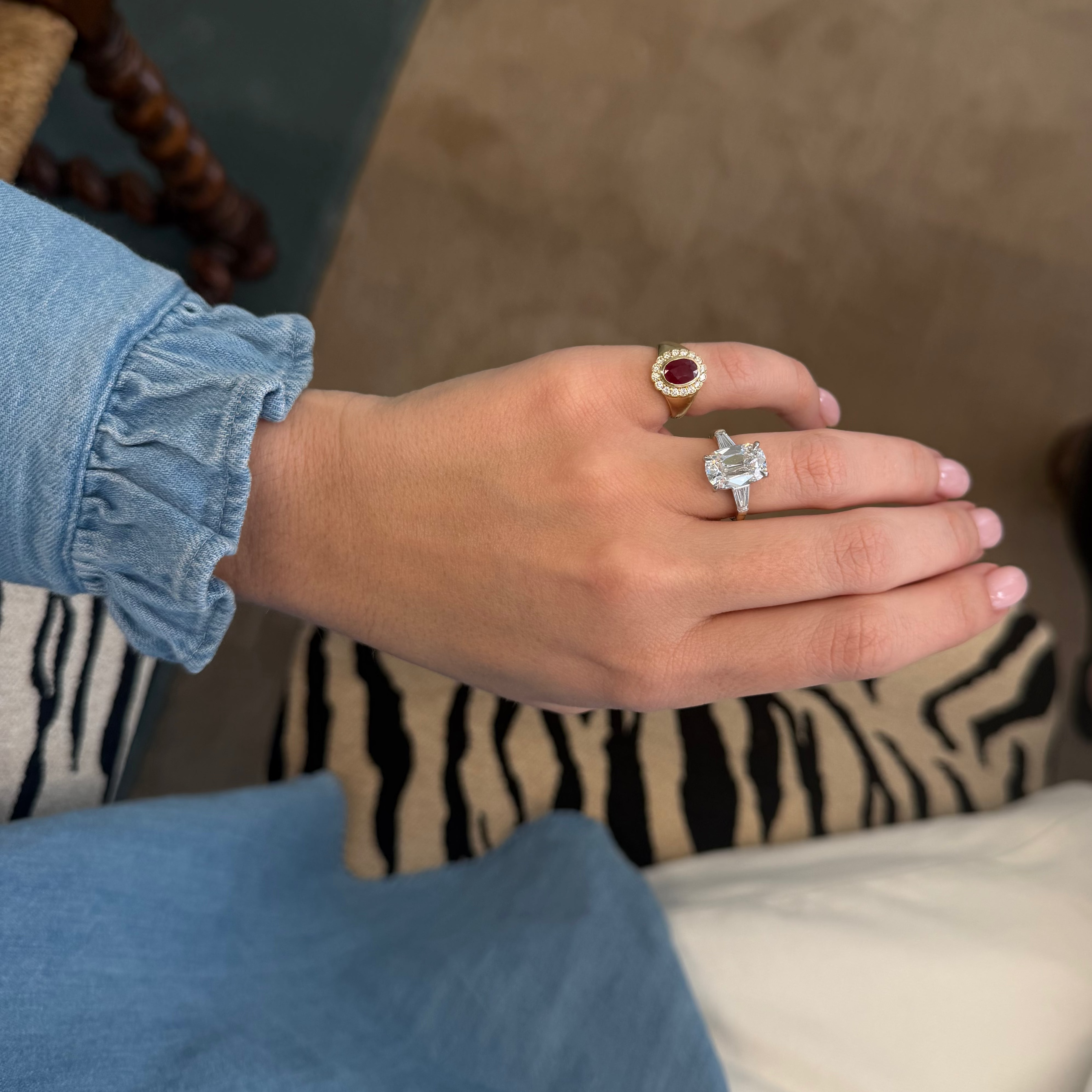 A person's hand wearing two rings: a gold ring with a round red gemstone and a silver ring with a square clear gemstone, shown in a home setting.