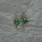 A pair of gold drop earrings featuring oval green emerald-cut stones and small clear crystal clusters, displayed on a textured gray paper background.