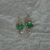 A pair of gold drop earrings featuring oval green emerald-cut stones and small clear crystal clusters, displayed on a textured gray paper background.