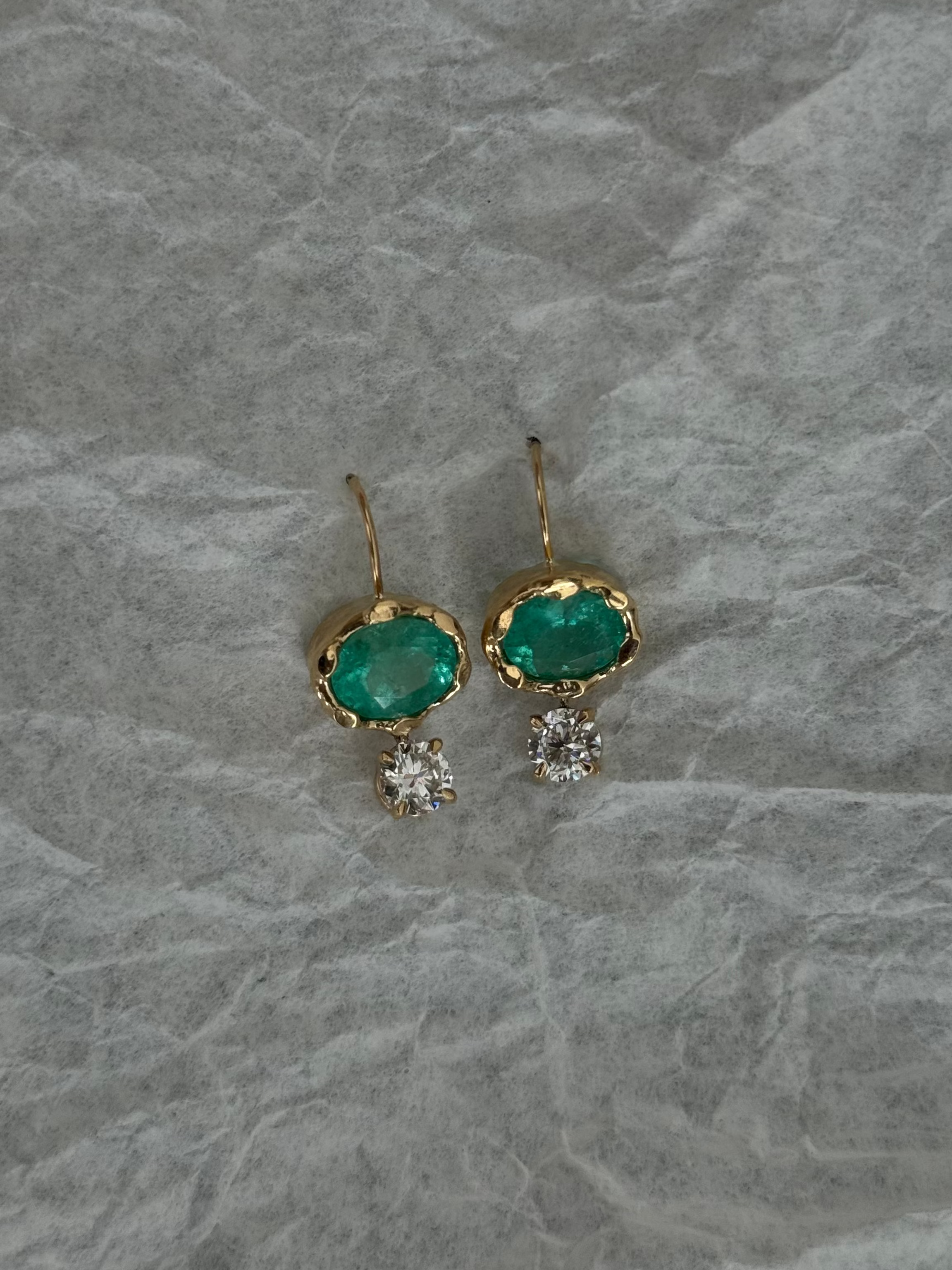 A pair of gold drop earrings featuring oval green emerald-cut stones and small clear crystal clusters, displayed on a textured gray paper background.
