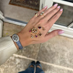 A hand with pink manicured nails displays a collection of gold rings and gemstones, worn with a blue strap watch, in front of a glass door.