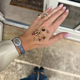A hand with pink manicured nails displays a collection of gold rings and gemstones, worn with a blue strap watch, in front of a glass door.