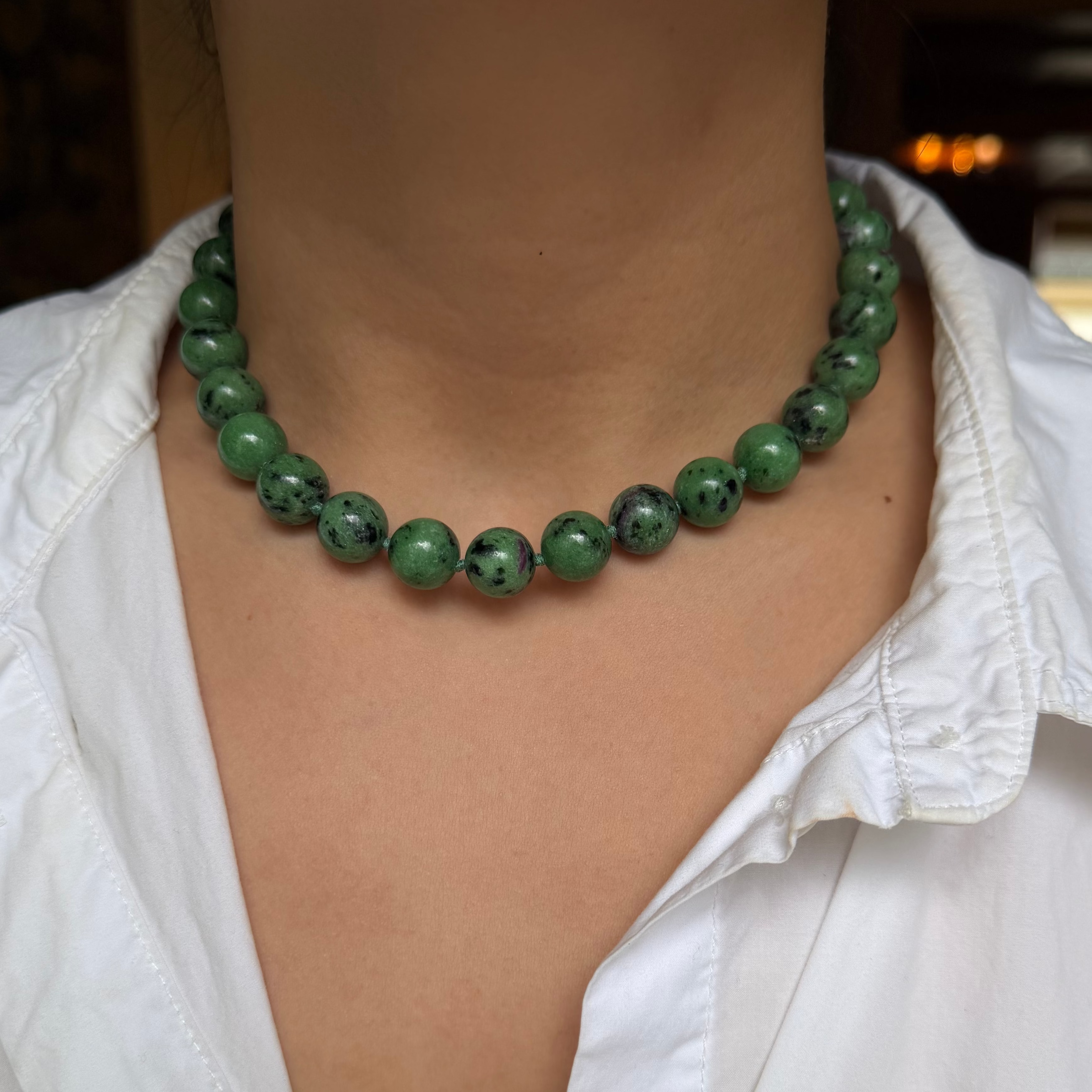 A close-up of a person wearing a green beaded necklace with dark marbled spots, styled with a white collared shirt.