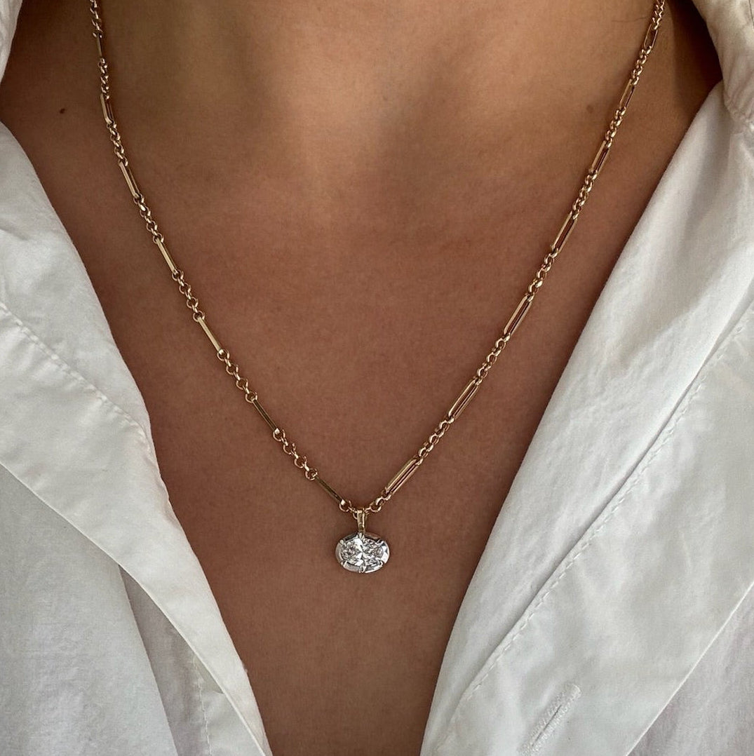 A close-up of a person wearing a delicate gold chain necklace with a small clear oval-shaped pendant, styled with a white collared shirt.