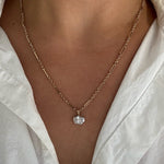 A close-up of a person wearing a delicate gold chain necklace with a small clear oval-shaped pendant, styled with a white collared shirt.