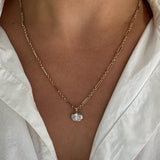 A close-up of a person wearing a delicate gold chain necklace with a small clear oval-shaped pendant, styled with a white collared shirt.