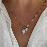 A close-up of a person wearing a delicate gold chain necklace with one teardrop-shaped clear gemstone pendant and one oval-shaped clear gemstone pendant, styled with a white collared shirt.