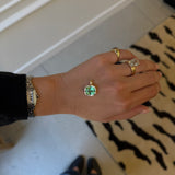 A gold pendant with a round green gemstone is shown on a person's hand, alongside other gold rings and a watch, in an indoor setting.