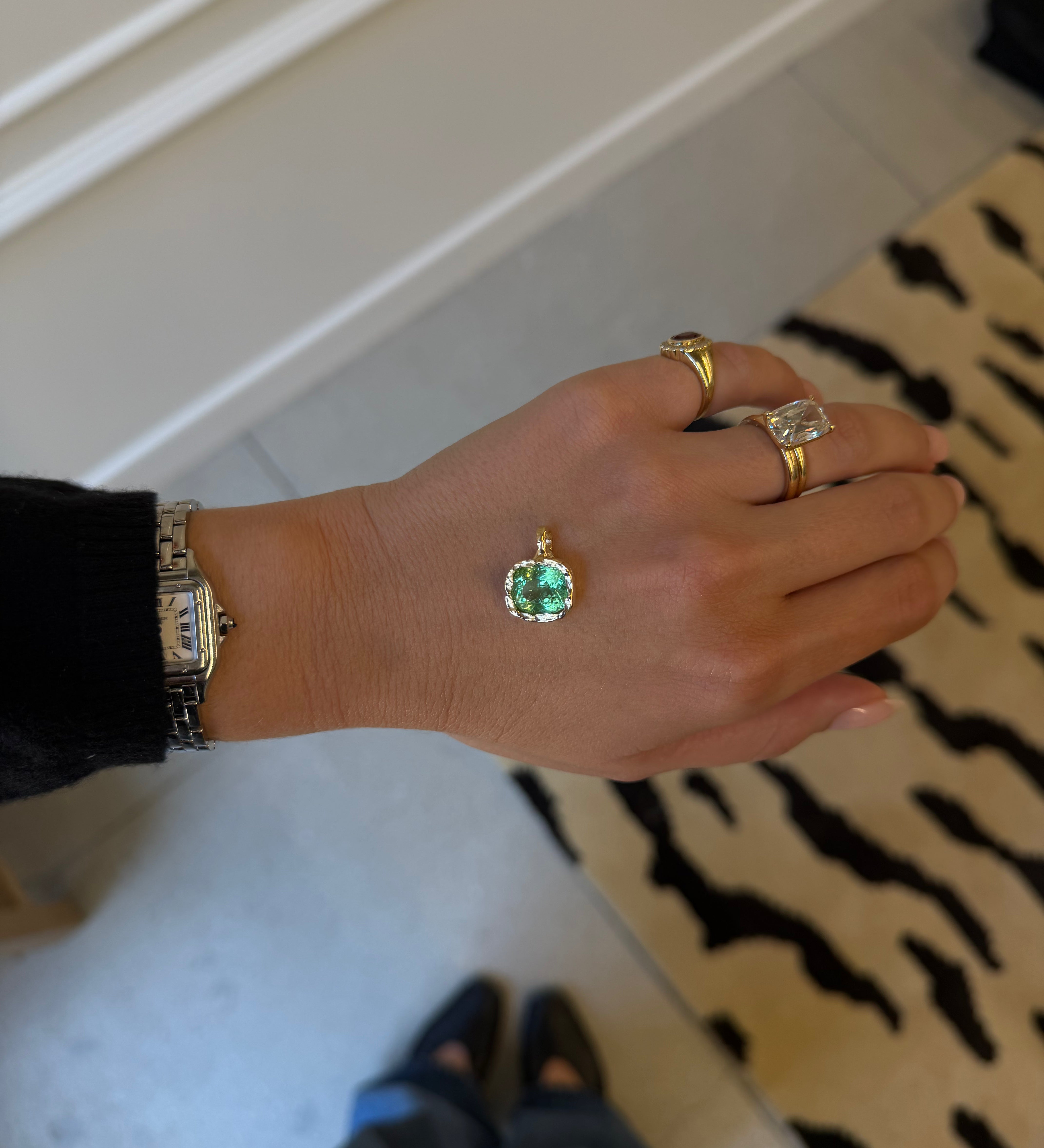 A gold pendant with a round green gemstone is shown on a person's hand, alongside other gold rings and a watch, in an indoor setting.