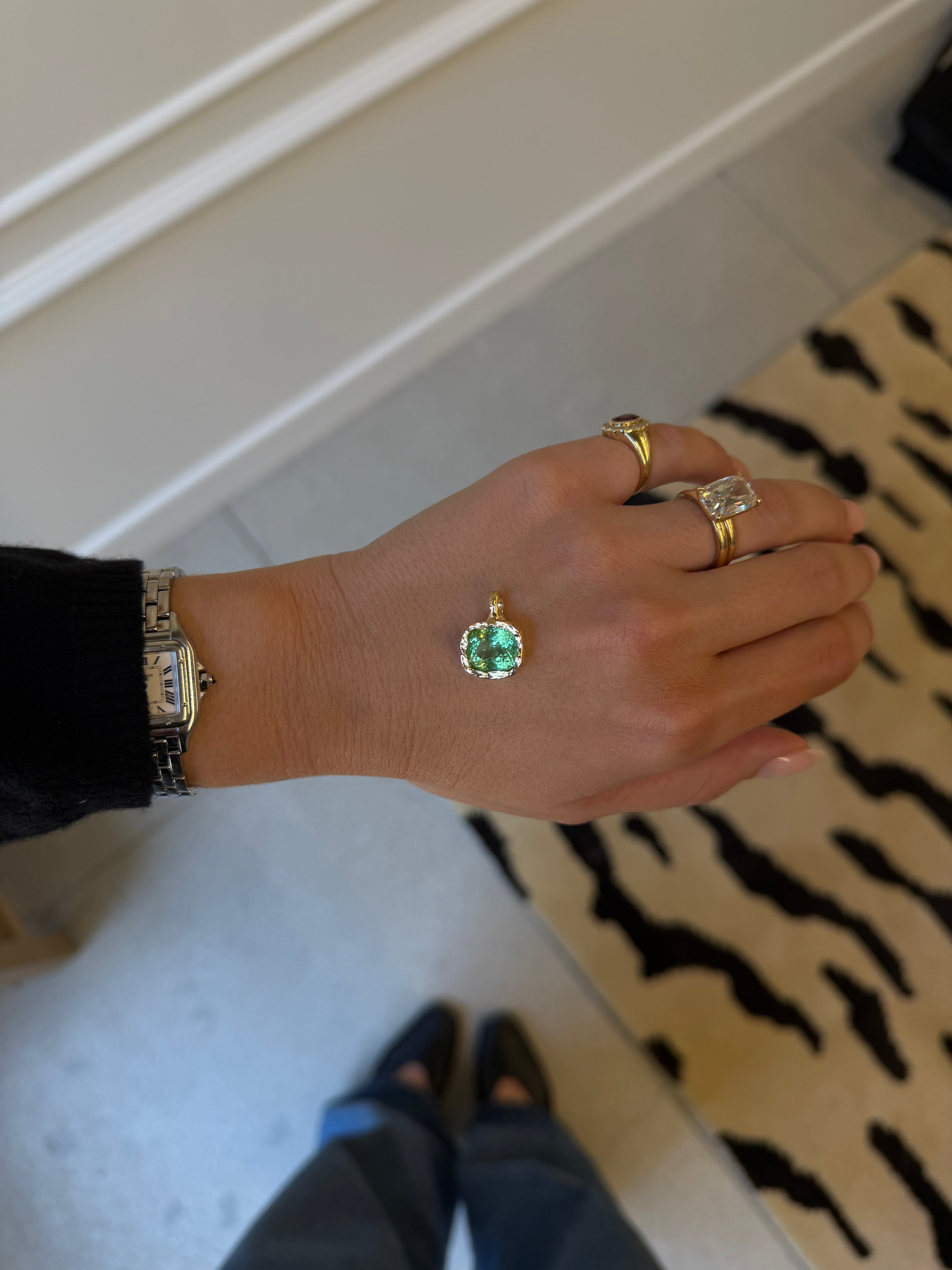 A gold pendant with a round green gemstone is shown on a person's hand, alongside other gold rings and a watch, in an indoor setting.