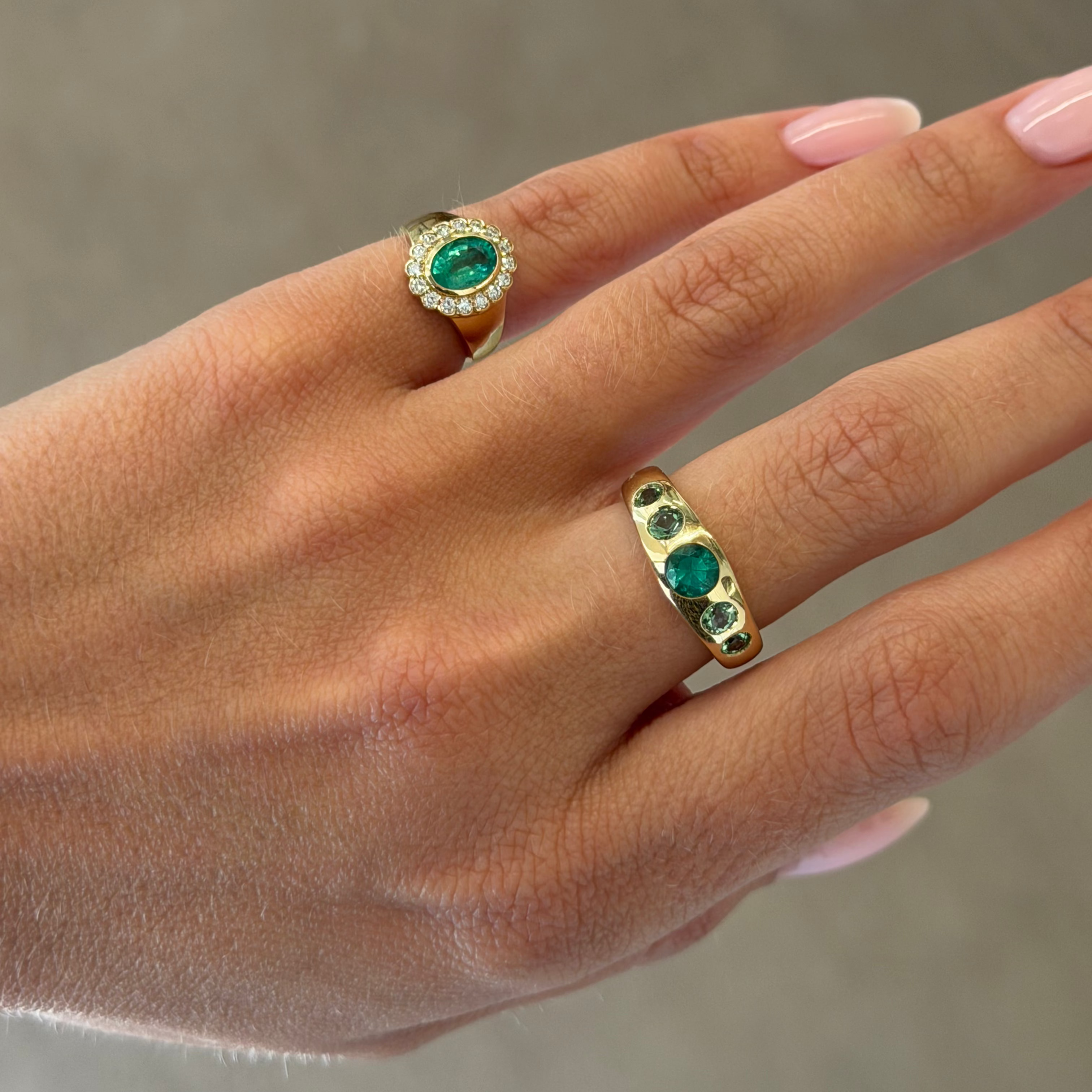 A close-up of a hand wearing two gold rings with green emerald-cut stones, set against a neutral gray background.