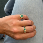 A close-up of a person's hand wearing two gold rings with green emerald-cut stones, resting on light blue denim fabric.
