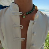 A close-up lifestyle shot of a person wearing a cream-colored Chinese-style jacket with toggle closures and a green beaded choker necklace with a green gemstone pendant, set against an outdoor beach background.