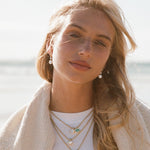 A woman with blonde hair wearing layered gold necklaces with small green and white charms, a white t-shirt, and a cream knit cardigan, set against a bright, sunlit beach background.
