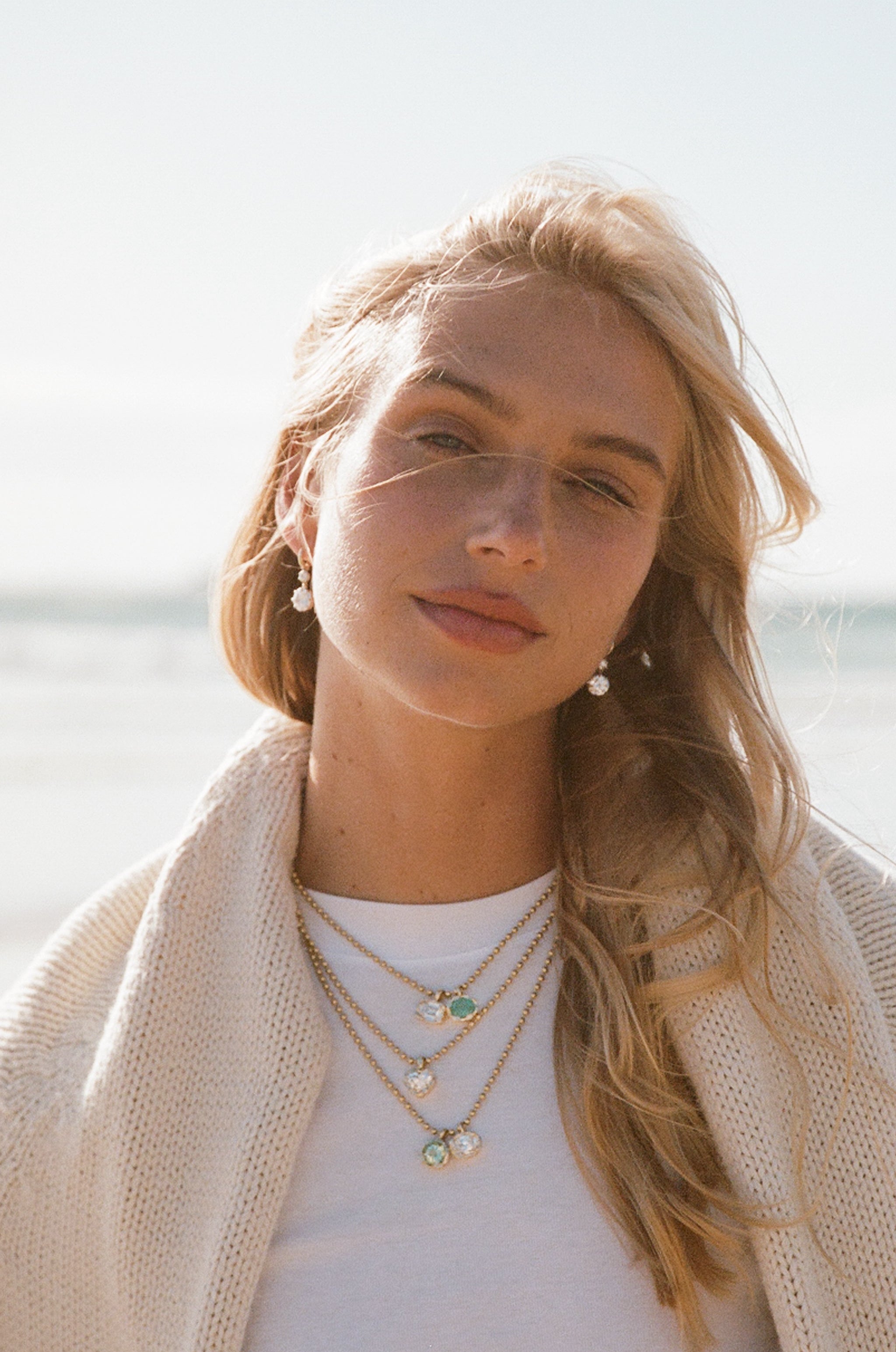 A woman with blonde hair wearing layered gold necklaces with small green and white charms, a white t-shirt, and a cream knit cardigan, set against a bright, sunlit beach background.