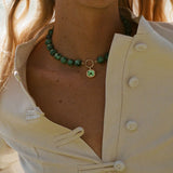 A close-up of a model wearing a green beaded choker necklace with a small gold ring and green square pendant, paired with a cream-colored button-up blouse.