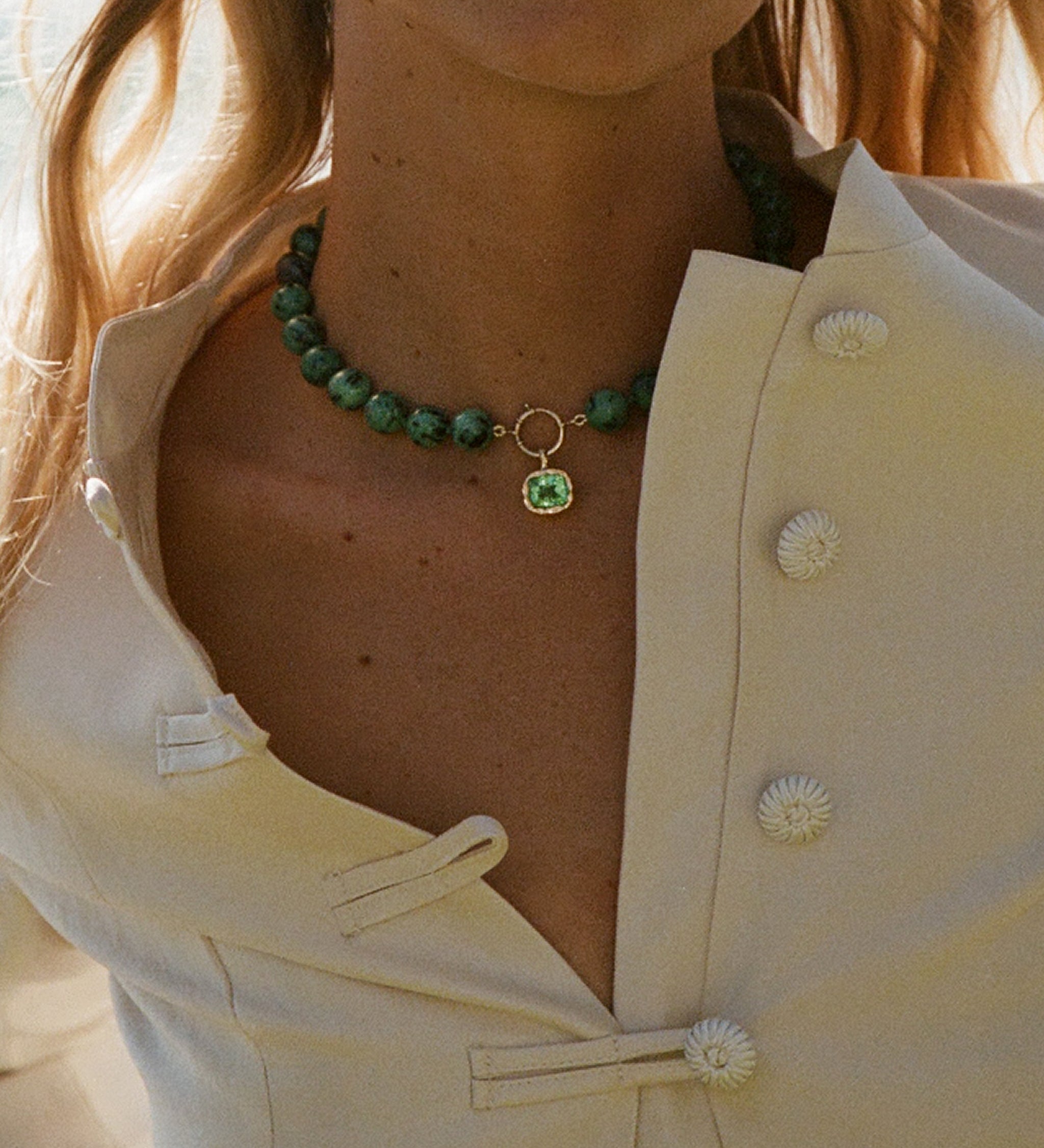 A close-up of a model wearing a green beaded choker necklace with a small gold ring and green square pendant, paired with a cream-colored button-up blouse.