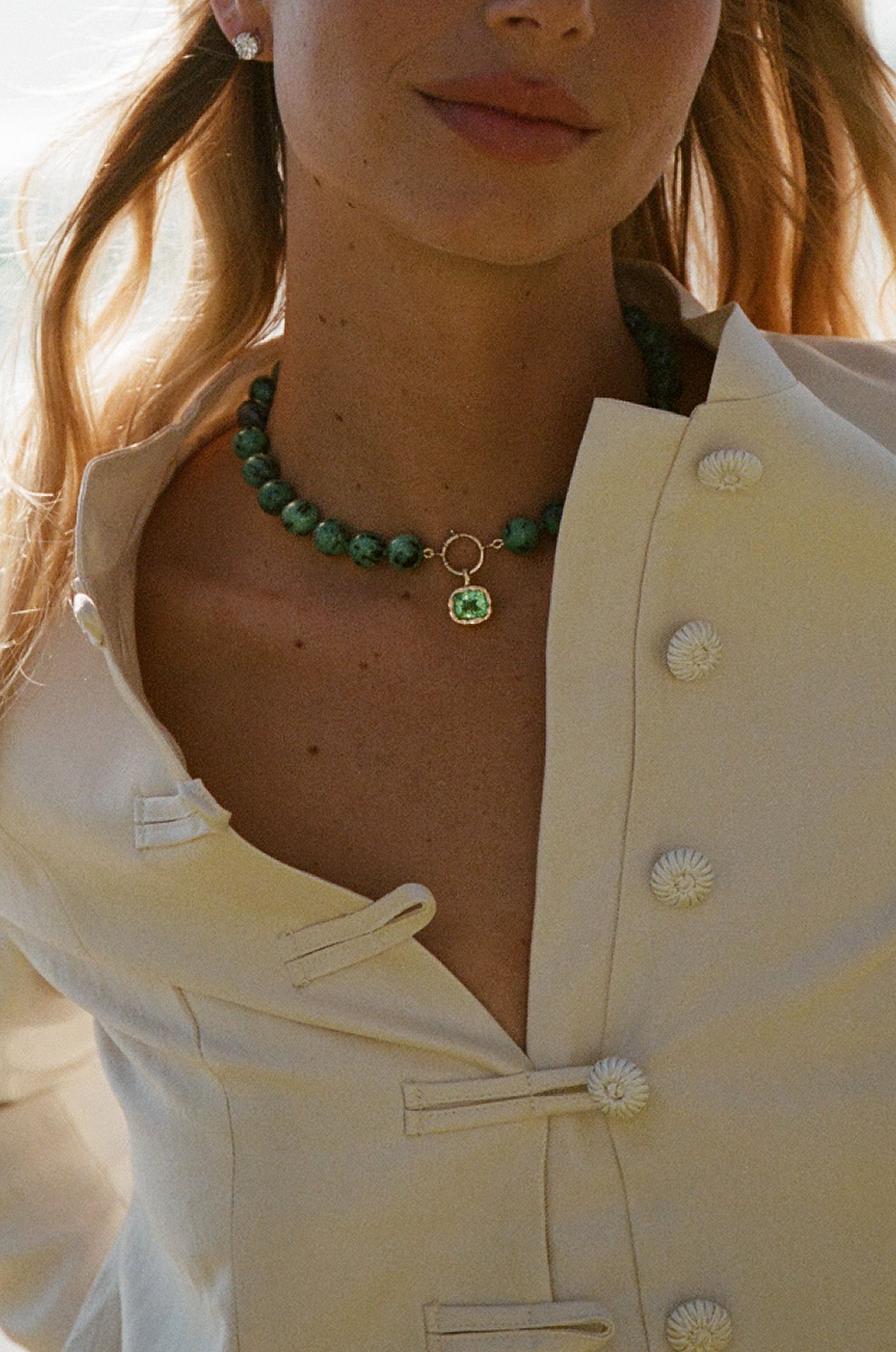A close-up of a model wearing a green beaded choker necklace with a small gold ring and green square pendant, paired with a cream-colored button-up blouse.