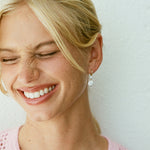 A close-up of a smiling blonde woman wearing a small silver hoop earring with a dangling clear crystal flower-like charm, against a plain light wall.